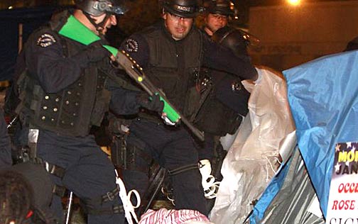 During the Nov. 30, 2011 raid on the Occupy L.A. camp, an LAPD officer inspects the inside of a tent with a "less than lethal" 12 gauge shotgun. Identified by its bright green stock and fore-grips, the weapon fires "bean bag" munitions, oblong cloth bags filled with up to 56 grams of lead pellets. The incapacitating impact of these rounds produces intense pain. Photo AP/Mark Boster/Pool 