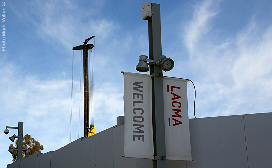 "LACMA Idyll." A formidable grey security wall some 15 ft tall, encircles what is left of museum grounds. The barrier completely forbids the public a view of ongoing construction. An incongruous "welcome" sign cloaks a drab tableau of destruction. Photo Mark Vallen ©. Feb 14 2021.